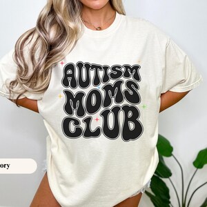 Puede incluir: Camiseta de color marfil con el texto "AUTISM MOMS CLUB" en una fuente retro negra. La camiseta tiene cuello redondo y mangas cortas. El texto está delineado en negro con acentos de estrellas coloridas.