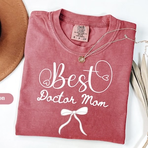Puede incluir: Camiseta de color carmesí con las palabras "Best Doctor Mom" en escritura blanca. Un collar plateado con un colgante descansa sobre la camiseta. La camiseta tiene un lazo gráfico y está doblada. Un sombrero marrón es visible en el fondo.