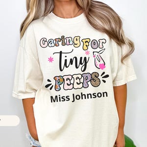 Puede incluir: Camiseta de color marfil con el texto "Caring for Tiny Peeps" en una fuente juguetona, con un conejito y detalles florales. El nombre "Miss Johnson" está impreso debajo. La camiseta tiene un corte holgado e informal.