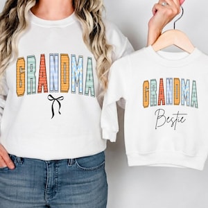Könnte beinhalten: Zwei weiße Langarm-Sweatshirts. Das größere zeigt das Wort GRANDMA in bunten, gemusterten Blockbuchstaben mit einer schwarzen Schleifengrafik darunter. Das kleinere zeigt GRANDMA in ähnlichen Buchstaben mit dem Wort Bestie in schwarzer Schrift.
