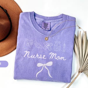 Puede incluir: Una camiseta violeta Comfort Colors con un gráfico blanco de símbolos médicos y las palabras "Nurse Mom". Un collar dorado y un sombrero marrón también están en la imagen.