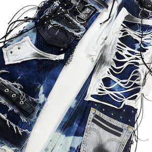 May include: Close-up of deconstructed denim jeans with distressed details. The jeans feature patches of dark blue denim, white fabric, and black mesh. There are also leather accents, grommets, and a zipper pocket. The design has a punk rock aesthetic.