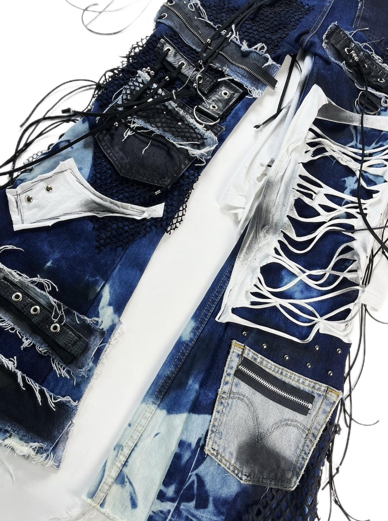 May include: Close-up of deconstructed denim jeans with distressed details. The jeans feature patches of dark blue denim, white fabric, and black mesh. There are also leather accents, grommets, and a zipper pocket. The design has a punk rock aesthetic.