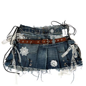 May include: A pleated blue denim mini skirt featuring a brown leather belt, silver metal grommets, white floral lace appliqués, and black cord lacing details. The hem is frayed and distressed, and a small white tag labeled RAGS is attached.