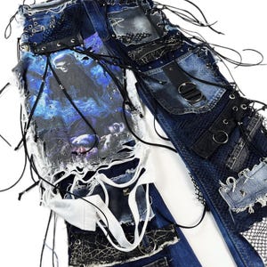 May include: Distressed denim jeans with multiple patches, lace, and grommet details. The jeans feature a graphic print on the front and are embellished with black straps and metal hardware. The design has a punk rock aesthetic.