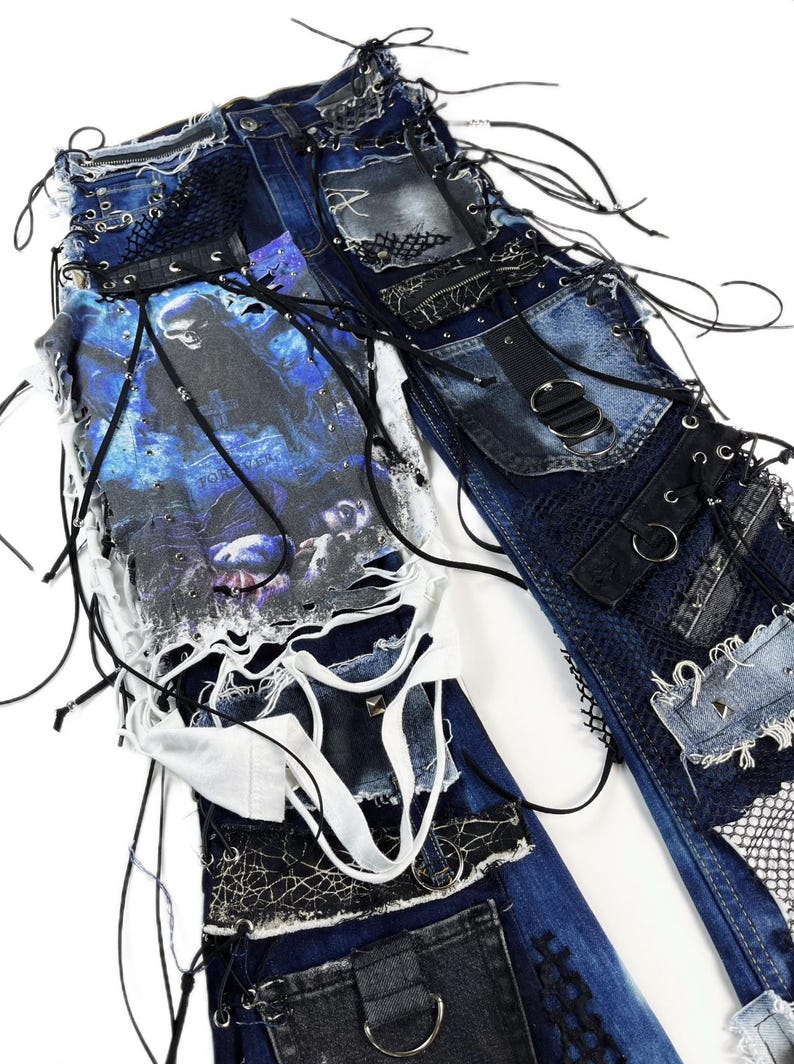 May include: Distressed denim jeans with multiple patches, lace, and grommet details. The jeans feature a graphic print on the front and are embellished with black straps and metal hardware. The design has a punk rock aesthetic.