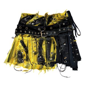 May include: A pleated skirt featuring a black and yellow distressed pattern with frayed edges. The garment is adorned with numerous silver safety pins, metal eyelets, and pyramid studs, and includes a black leather belt with a silver buckle.