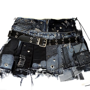 May include: A pleated mini skirt constructed from distressed blue denim and black fabric panels. The skirt features a black leather belt with metal grommets, silver safety pins, metal studs, and black mesh overlays with frayed edges.