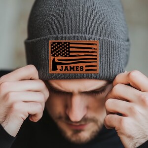May include: A gray knit beanie with a brown leather patch featuring an American flag, an axe, and the name "JAMES".