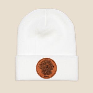 May include: White knit beanie with a brown leather patch featuring a black and white illustration of a dog with the name "Coco" written below.