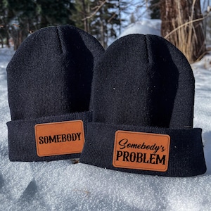 May include: Two black knit beanies with brown leather patches. The patch on the left beanie says "Somebody". The patch on the right beanie says "Somebody's Problem".