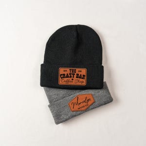 May include: Two beanies, one black and one gray, are displayed. The black beanie features a brown leather patch with the text "Crazy Bar Coffee Shop." The gray beanie has a brown leather patch with the name "Marcelyn Wood Co."