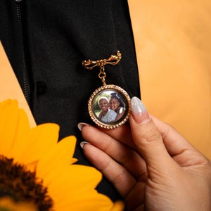 May include: A gold-colored brooch with a bow-shaped top and a dangling round pendant. The pendant features a photo of two people and is surrounded by small, clear crystals. The brooch is pinned to a black garment, with a yellow flower in the foreground.