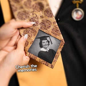 May include: A brown and gold patterned tie with a black and white photo of a woman wearing a graduation cap and gown. The photo is in a square frame and the text "Cherish the memories" is below the photo.