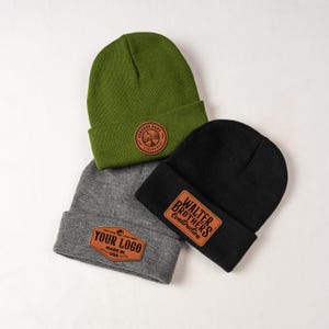 May include: Three knit beanies in green, gray, and black. Each beanie has a leather patch with a logo. The green beanie has a circular patch, the gray beanie has a rectangular patch with the text "YOUR LOGO", and the black beanie has a rectangular patch with the text "WALTER BROTHERS Construction".