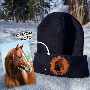 May include: Black knit beanie with a brown leather patch featuring a horse silhouette and the name "Bella".