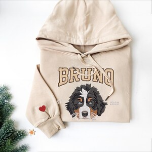 May include: Beige hoodie with a brown embroidered dog and the name "BRUNO" in gold lettering. The hoodie also has a red heart on the sleeve and the year "2023" embroidered on the bottom right.