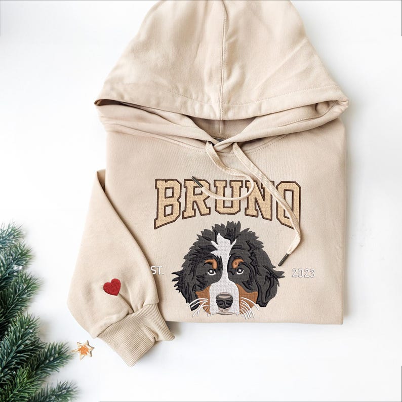 May include: Beige hoodie with a brown embroidered dog and the name "BRUNO" in gold lettering. The hoodie also has a red heart on the sleeve and the year "2023" embroidered on the bottom right.