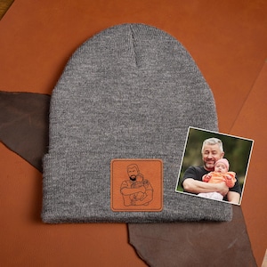 May include: Gray knitted beanie with a brown leather patch featuring a line drawing of a person holding two children. The patch is sewn onto the front of the beanie.