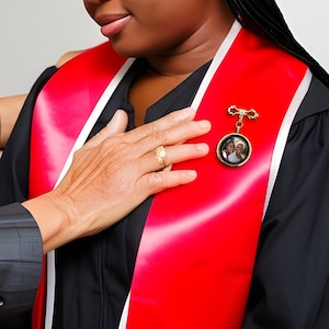 May include: A red and black graduation gown with a gold pin featuring a photo of two people. The pin is worn on the left side of the chest.