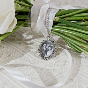 May include: A white bouquet of flowers with a silver locket charm attached. The locket contains a black and white photo of a couple.