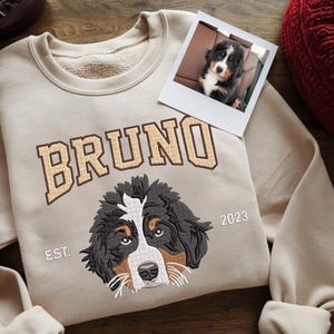 May include: A beige sweatshirt with a brown Bernese Mountain Dog embroidered on the front. The dog's name, "BRUNO", is embroidered in brown above the dog's image. The text "EST. 2023" is embroidered in white below the dog's image.
