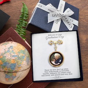 May include: A gold-colored graduation pin with a photo pendant, presented in a black box with the text "With you on your Graduation Day." The pin is displayed on a white card with a sentimental quote. A blue gift box with a white ribbon is also visible.