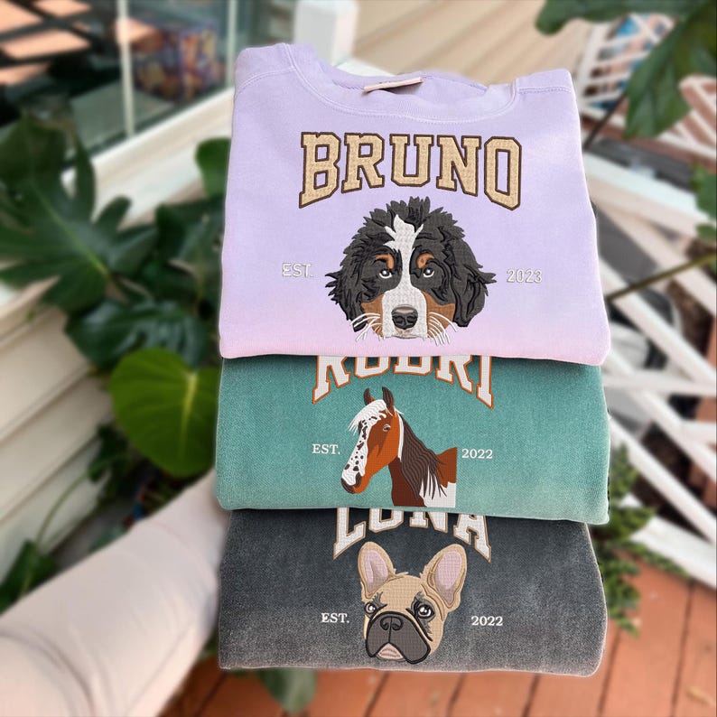 May include: Three sweatshirts stacked on top of each other. The top sweatshirt is light purple with the text "BRUNO" in gold letters and a Bernese Mountain Dog embroidered on the front. The middle sweatshirt is teal green with the text "ROOKIE" in gold letters and a horse embroidered on the front. The bottom sweatshirt is dark gray with the text "LUNA" in gold letters and a French Bulldog embroidered on the front.