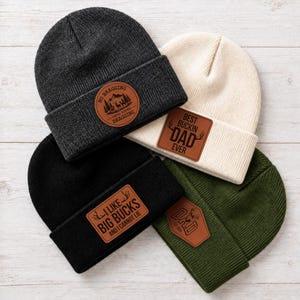 May include: Four knit beanies in charcoal, cream, black, and olive green. Each features a brown leather patch with engraved text: No Bragging Until You're Dragging, Best Buckin Dad Ever, I Like Big Bucks And I Cannot Lie, and Dad Est 2020.