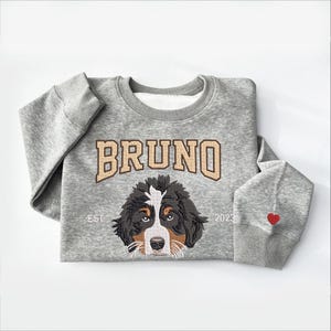 May include: A gray sweatshirt with a brown and black Bernese Mountain Dog embroidered on the front. The dog's name, "BRUNO", is embroidered above the dog in a brown and white font. The text "EST. 2023" is embroidered below the dog. A red heart is embroidered on the right sleeve.
