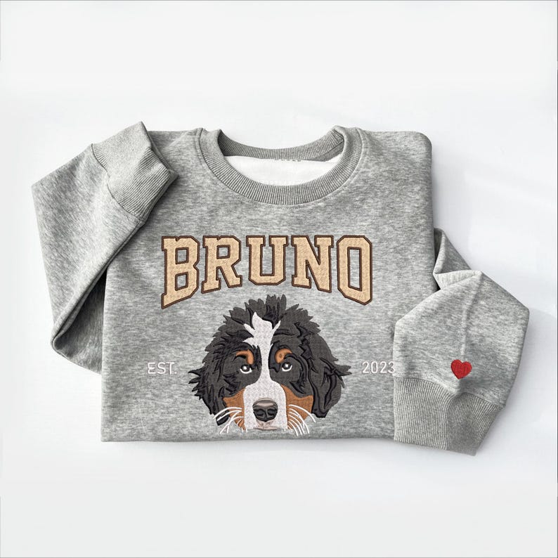 May include: A gray sweatshirt with a brown and black Bernese Mountain Dog embroidered on the front. The dog's name, "BRUNO", is embroidered above the dog in a brown and white font. The text "EST. 2023" is embroidered below the dog. A red heart is embroidered on the right sleeve.