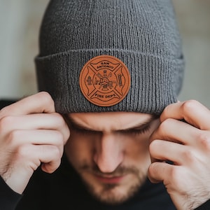 May include: A gray knit beanie with a brown leather patch featuring the San Antonio Fire Department logo. The logo is a circle with a cross inside, and it includes a fire hydrant, an axe, a ladder, and a firefighter's helmet.