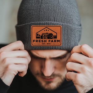 May include: A gray beanie with a brown leather patch featuring a black tractor and the text "FRESH FARM - EST. 1945 - "