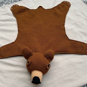 May include: A brown crocheted bear rug with a stitched nose and black eyes. The rug is on a light brown surface.