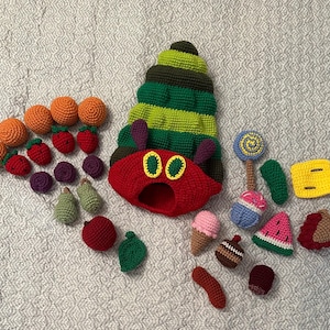 May include: A collection of colorful, crocheted food items and a caterpillar hat. The set includes oranges, strawberries, plums, pears, an apple, a lollipop, ice cream cones, a watermelon slice, and a cupcake. The caterpillar hat is red with green and yellow accents.
