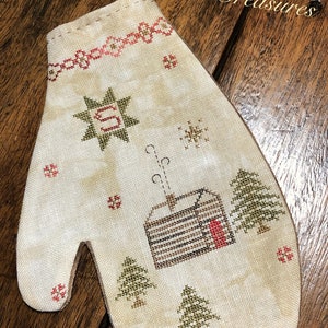May include: A white fabric mitten with a cross-stitch design featuring a cabin with smoke coming from the chimney, trees, and stars. The text "Dulaney Woods Treasures" is in the upper right corner.