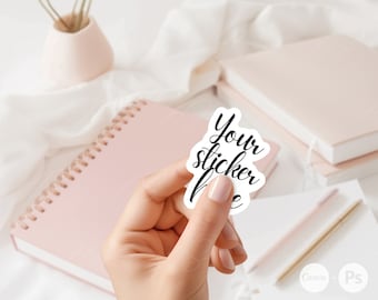 May include: A white sticker with black cursive text that reads "Your sticker here". The sticker is being held by a hand against a background of pink and white notebooks and a white surface.