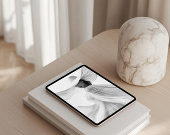 May include: A tablet computer displaying a black and white image of a person wearing a flowing garment. The tablet is resting on top of two white books on a light wood surface.