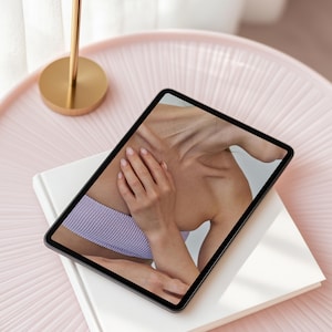 May include: A tablet computer displaying a close-up image of a person's shoulder and arm, wearing a light purple top. The tablet is lying on a white book, which is on a pink table.