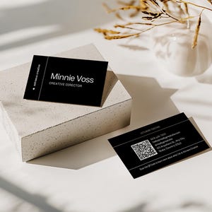 May include: Black business cards with white text, including the name "Minnie Voss, Creative Director." One card rests on a concrete block, another lies flat. The cards feature a QR code and contact information. The background is white with shadows.