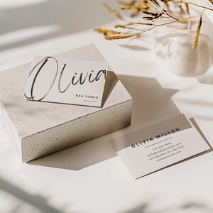 May include: Two business cards with the names "Olivia" and "Olivia Wilson" printed on them. The "Olivia" card is on a concrete block. The "Olivia" card says "Nail Studio". The "Olivia Wilson" card has contact information. A white vase and dried plants are in the background.