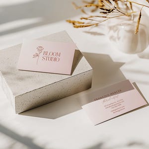 May include: Two business cards with a floral design and the text "BLOOM STUDIO" and "FRANCOIS MERCER Floral artist" are displayed on a white surface. The cards are light pink and brown. A white vase and dried plants are in the background.