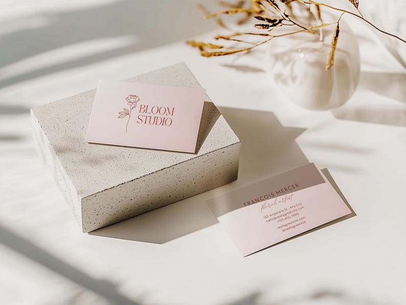 May include: Two business cards with a floral design and the text "BLOOM STUDIO" and "FRANCOIS MERCER Floral artist" are displayed on a white surface. The cards are light pink and brown. A white vase and dried plants are in the background.