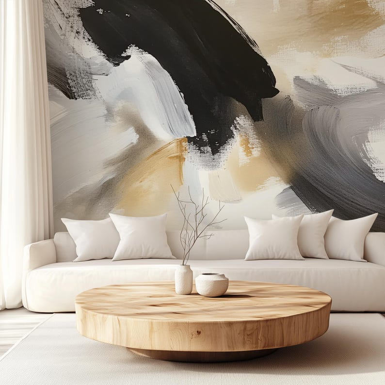 May include: A white sofa with white cushions sits in front of a wall with an abstract painting in shades of black, white, and gold. A round wooden coffee table sits in front of the sofa with a small white vase and a small grey bowl on top.