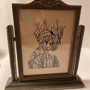 May include: A framed silhouette print of a person with branches and birds forming their head and body. The frame is dark gray with a distressed finish and has a stand.