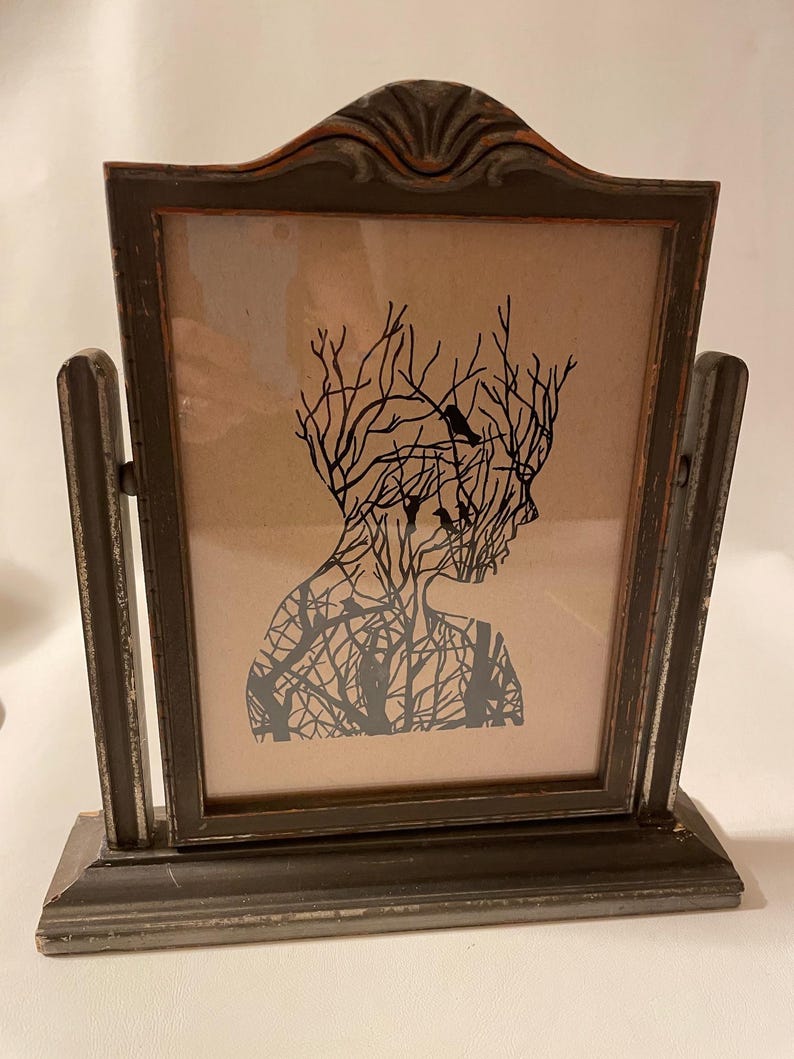 May include: A framed silhouette print of a person with branches and birds forming their head and body. The frame is dark gray with a distressed finish and has a stand.