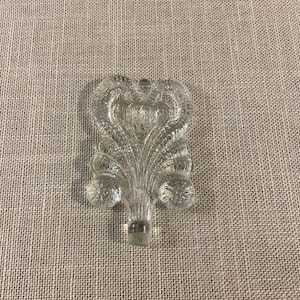 Puede incluir: Tapón de botella de vidrio transparente con un diseño ornamentado y estilizado. El tapón presenta un elemento central vertical con una serie de formas curvas en forma de hoja y acentos redondeados. La base del tapón tiene forma cilíndrica.