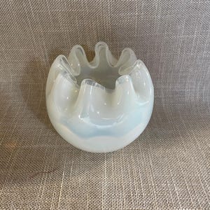 May include: A white, ruffled glass bowl with a rounded shape. The bowl has a milky, opalescent appearance and a wavy, scalloped rim. The bowl is sitting on a textured, neutral-colored surface.