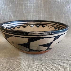 Puede incluir: Cuenco de cerámica pintado a mano con interior y exterior color crema. El cuenco presenta un diseño geométrico negro de triángulos y diamantes. El borde es negro con triángulos blancos y negros. La base es de color marrón rojizo.