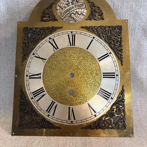 May include: A brass clock face featuring a central gold textured circle with black Roman numerals on a silver ring. The top arch displays a globe graphic with the text Tempus Fugit. Ornate dark metal corner spandrels decorate the rectangular plate.